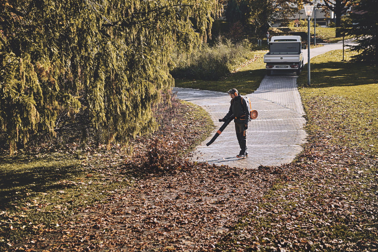 A man in work equipment using a cordless leaf blower to remove leaves from a park path