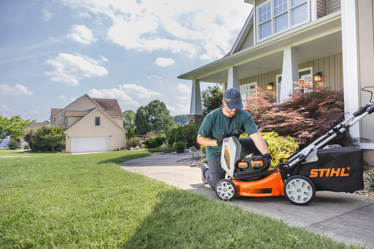 A man is halfway through mowing untidy and overgrown grass using a STIHL RMA 448 P-C cordless lawn mower, in a garden surrounded by tall shrubs and plants